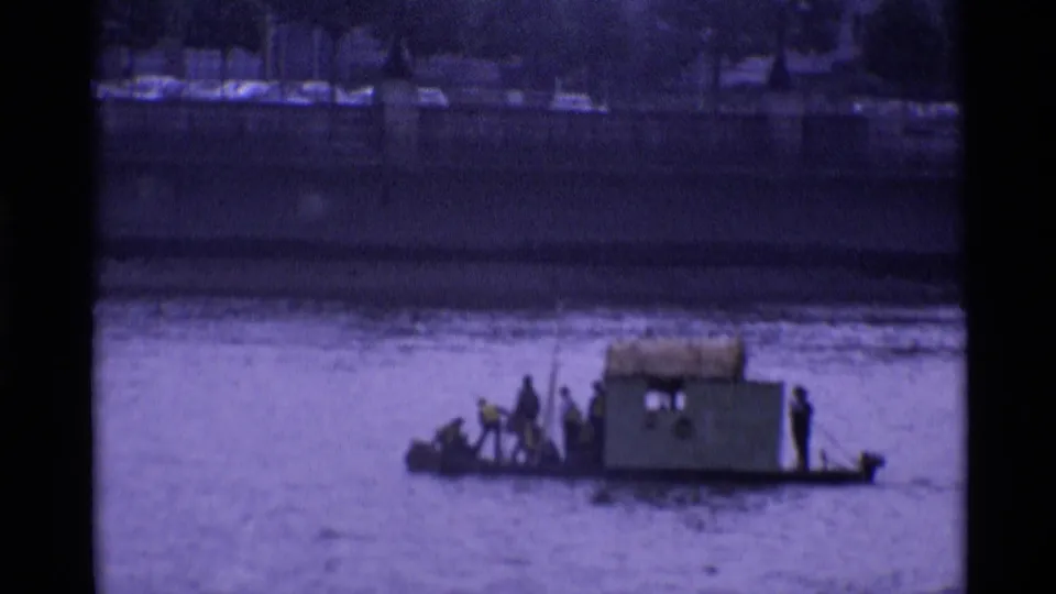 Small boat carrying distant people crosses the London river view, with dark pillar edges and embankment in the background.