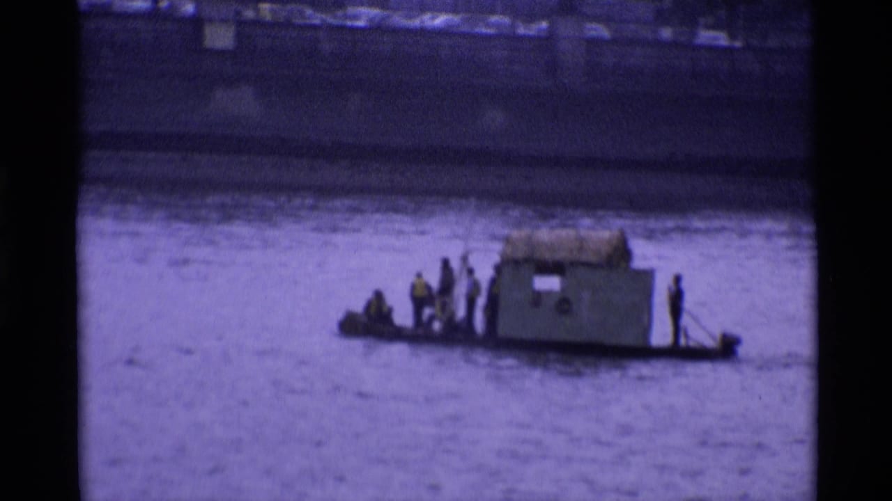 Men stand and sit on a low boat with a small shelter on the river.