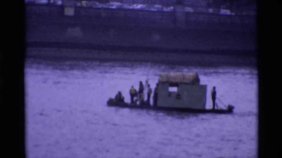 Distant 1969 London river view of men gathered around a small cabin-like structure on a makeshift boat.