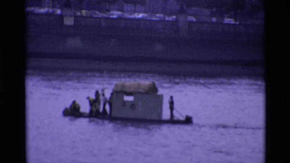 1969 London home-movie frame of a low improvised river craft carrying several men past a distant bank.