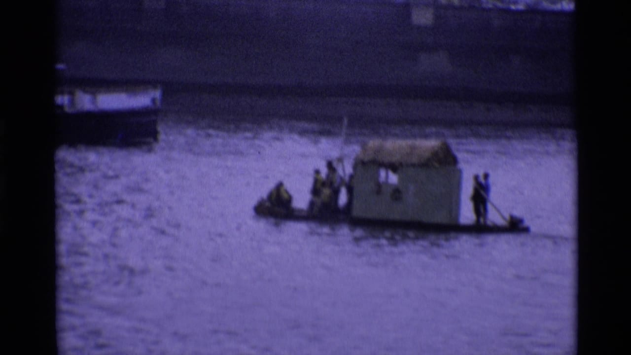 1969 London home-movie frame showing a makeshift cabin boat or raft with people on a broad urban river.