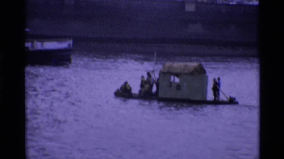 1969 London home-movie frame showing a makeshift cabin boat or raft with people on a broad urban river.