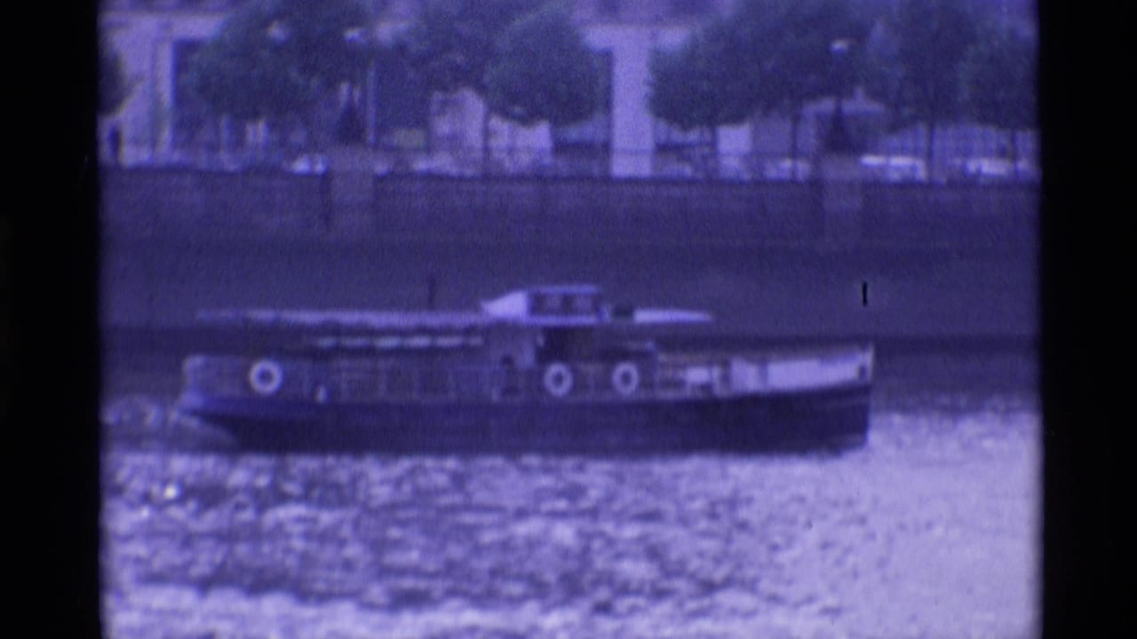 1969 London waterfront frame with a passenger tour boat passing along the river before trees and city buildings.
