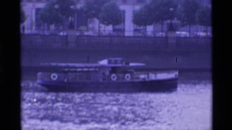 1969 London waterfront frame with a passenger tour boat passing along the river before trees and city buildings.