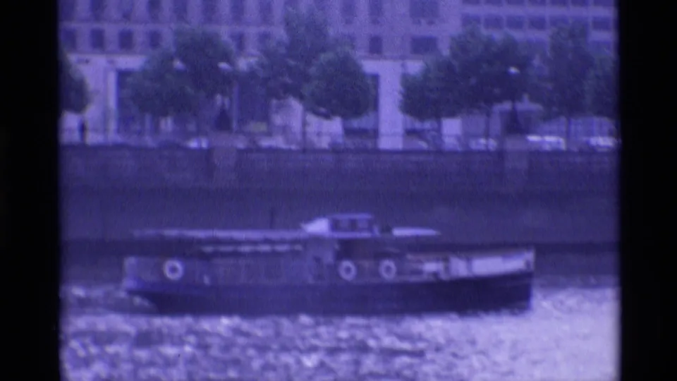 Vintage 8mm London frame showing a tour boat crossing rippled river water below an urban embankment.