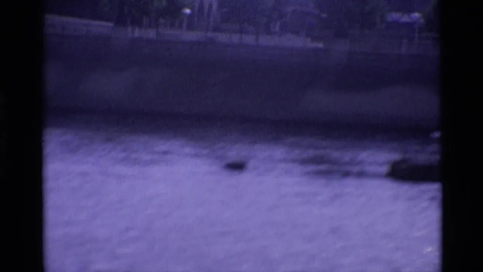 Soft, handheld 1969 London river view with water, embankment shadow, and partial dark boat edge at the frame side.