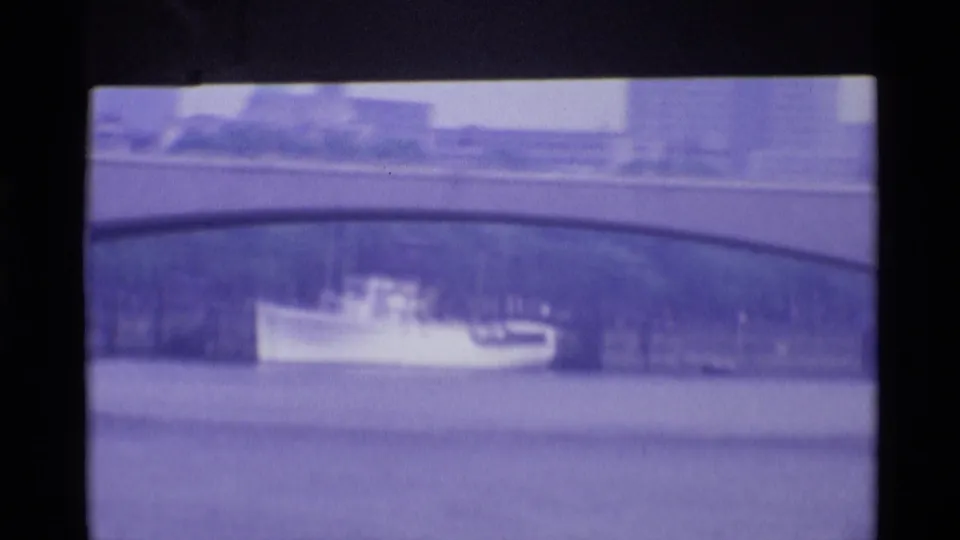 1969 London river view with a small boat passing beneath or near a stone bridge, framed by distant waterfront buildings.