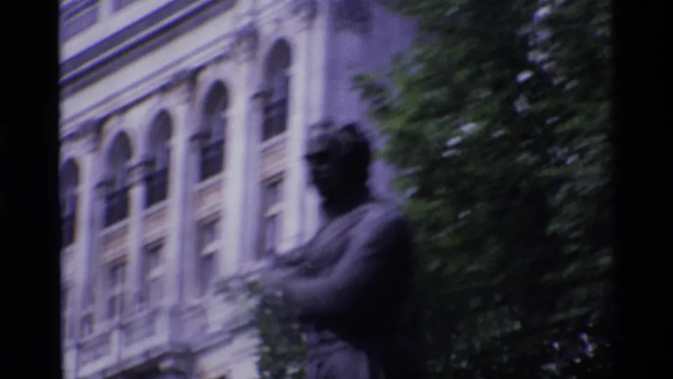 1969 London visitor-shot frame of a dark statue near trees, with a large building facade and arched windows behind it.