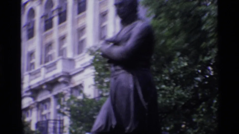 1969 London 8mm frame showing a dark public statue near leafy trees and a pale, ornate building facade.