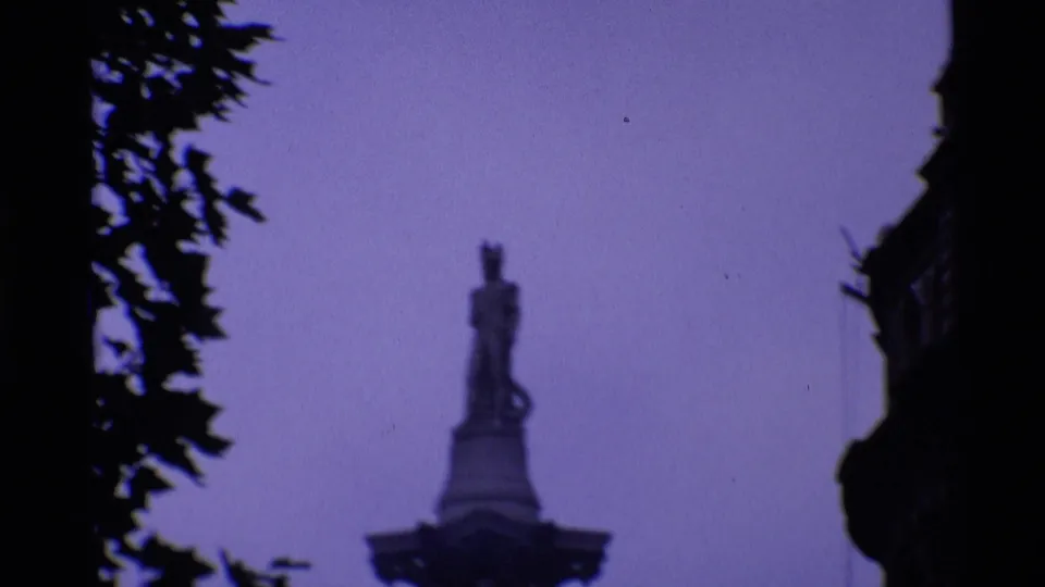 1969 London 8mm frame showing a distant statue-topped monument against pale sky, framed by tree leaves and dark building edges.