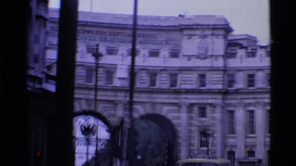 1969 London home-movie frame showing a formal stone building near Trafalgar Square, partly obscured by trees and street fixtures.