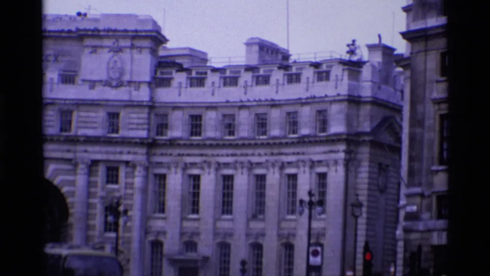 Handheld 1969 London view of a curved stone facade near Trafalgar Square with street lamps, trees, and distant street activity.