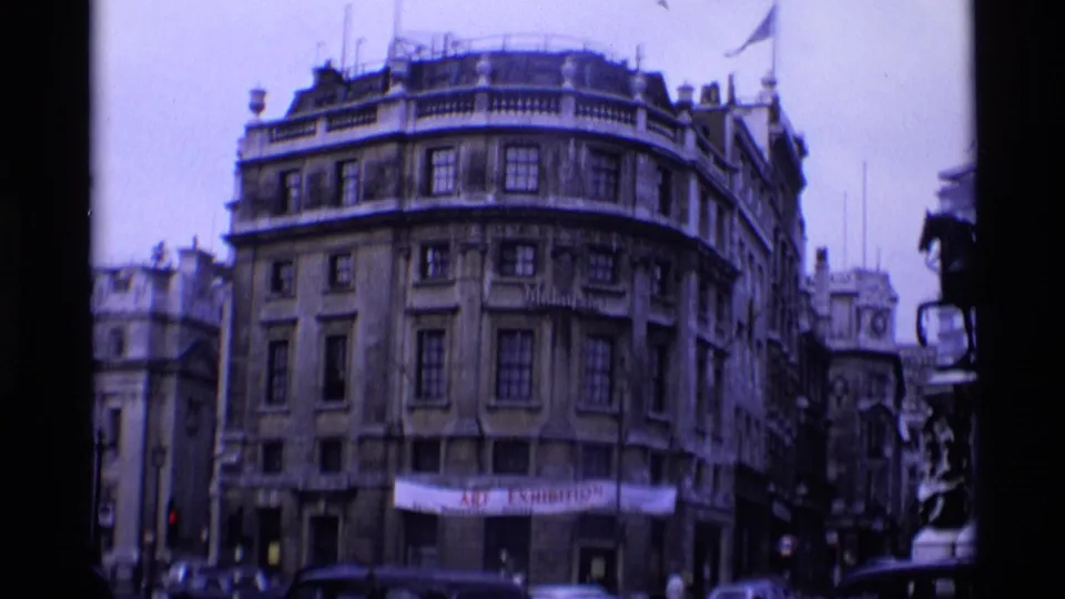 1969 London corner street scene showing a multi-story historic building, traffic, and a street-level banner.