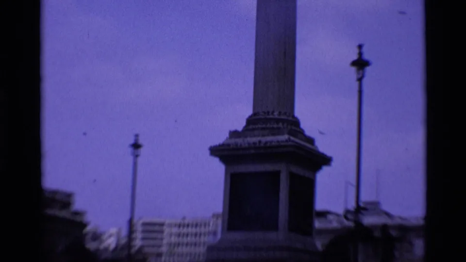 1969 London 8mm frame looking upward at Nelson's Column, its base, nearby lamp posts, and surrounding city buildings.