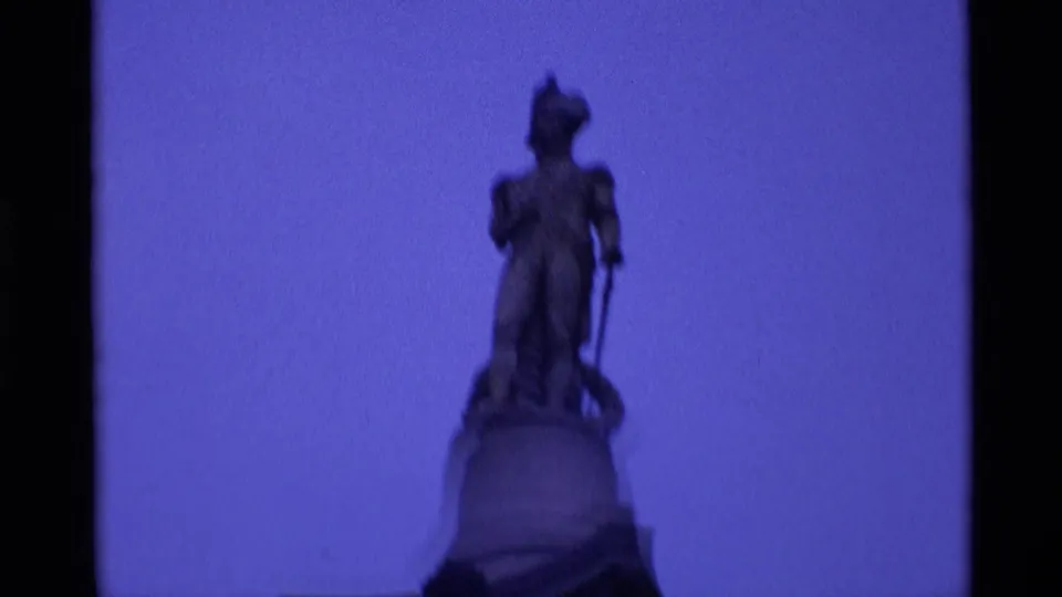 1969 London 8mm frame showing a low-angle view of a robed statue on a pedestal, silhouetted against blue sky.