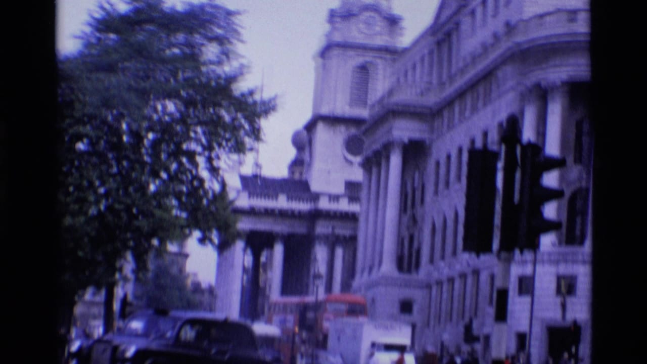 1969 London 8mm frame showing a broad street view with ornate stone buildings, trees, traffic lights, cars, and a red double-decker bus.