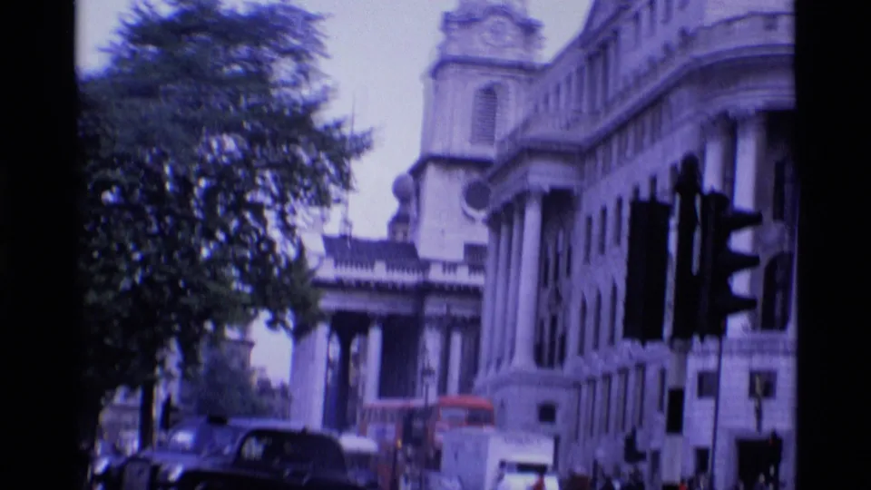1969 London 8mm frame showing a broad street view with ornate stone buildings, trees, traffic lights, cars, and a red double-decker bus.