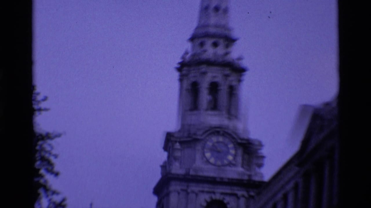 1969 London 8mm frame looking up at a church clock tower beside a darker roofline under a purple color cast.