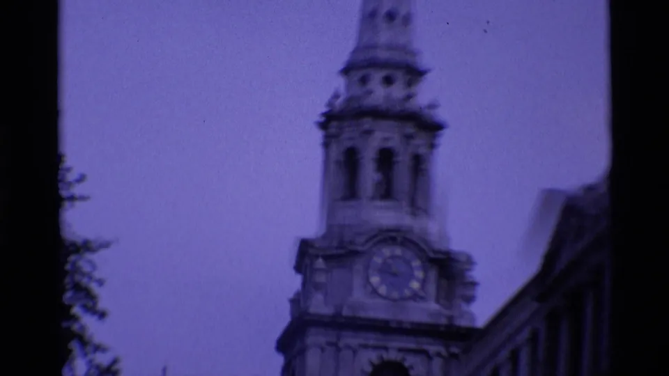 1969 London 8mm frame looking up at a church clock tower beside a darker roofline under a purple color cast.