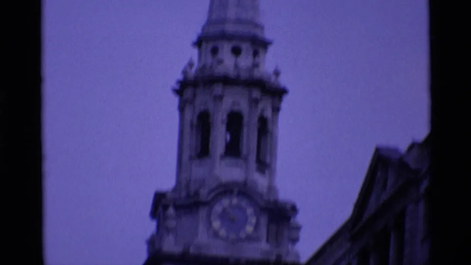 1969 London 8mm close view of a stone church clock tower with arched openings, clock face, and adjacent roofline.