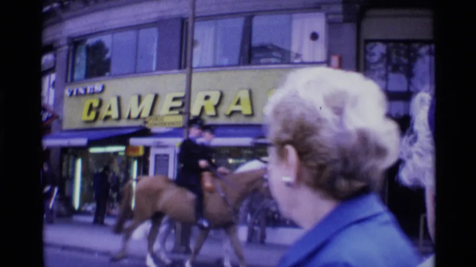 Mounted police officers and horses pass in front of a London camera-shop storefront while pedestrians watch in 1969 home-movie footage.