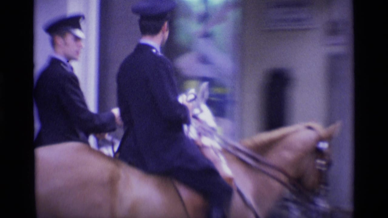 Two uniformed men in dark jackets and white hats ride horses through a London street in 1969 footage.