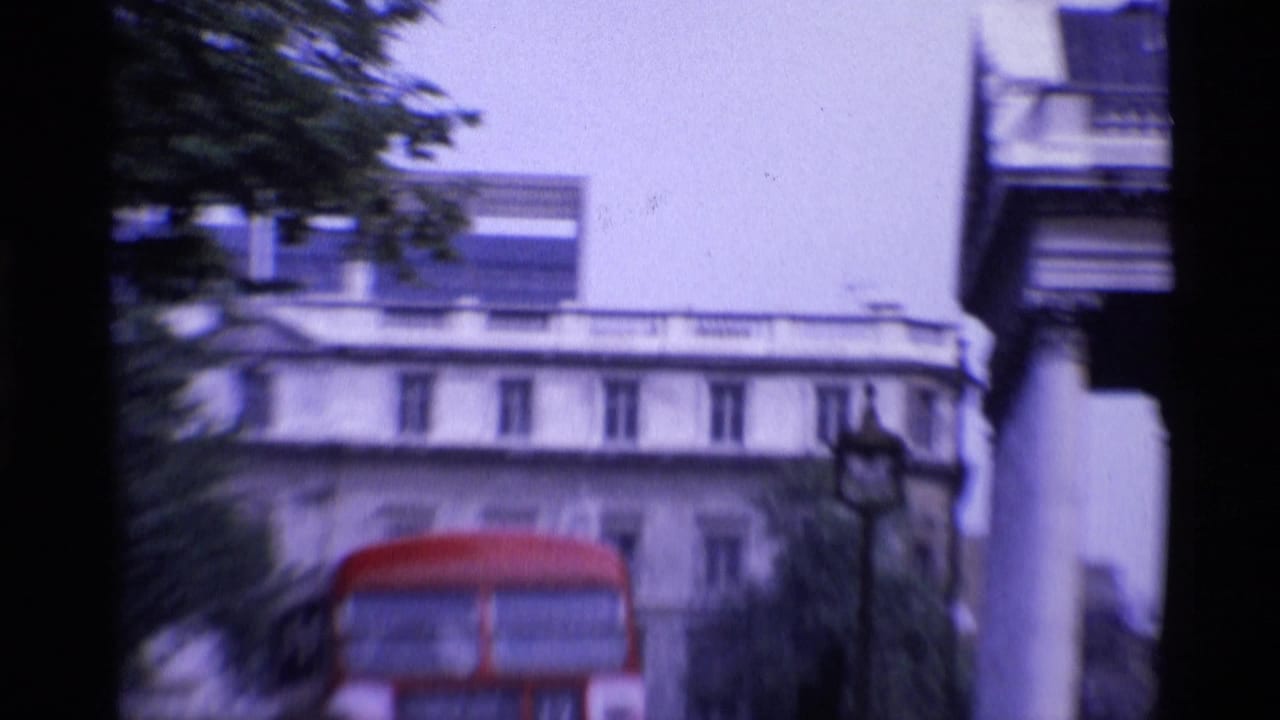 Red double-decker bus passes between large city buildings.