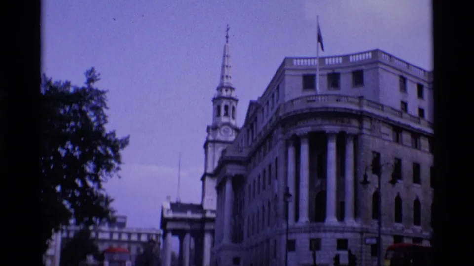 A London street view frames a grand stone building, a church spire, trees, and part of a red bus in 1969 home-movie footage.