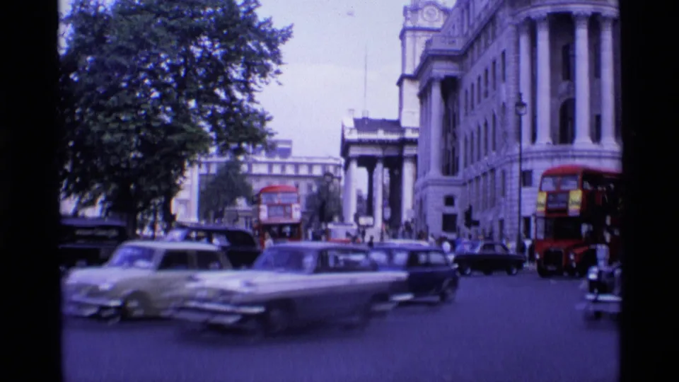 1969 London street view with red double-decker buses, vintage cars, a black taxi, pedestrians, trees, and a grand stone building.