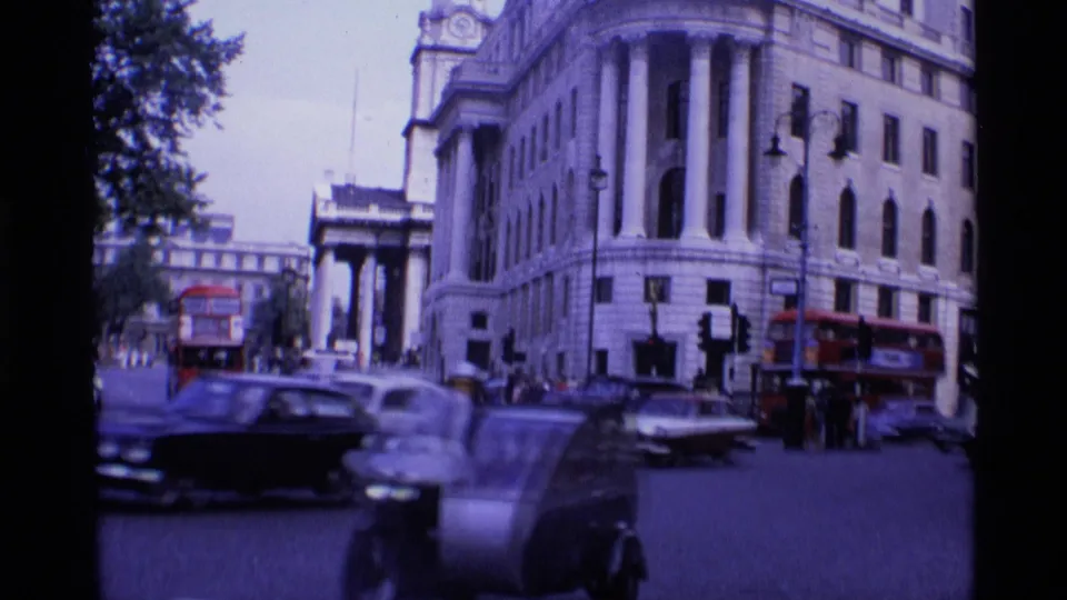Handheld 1969 London street footage showing motion-blurred cars, a red bus, traffic lights, pedestrians, and formal stone architecture.