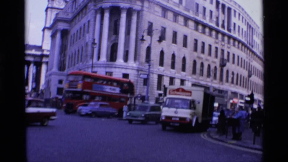 1969 London intersection with a red double-decker bus, white delivery truck, vintage cars, pedestrians, and a long stone building frontage.