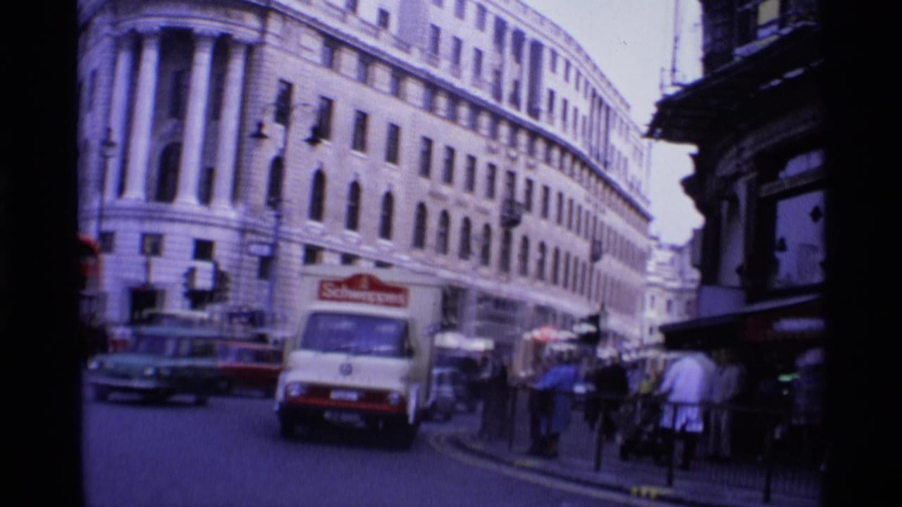 1969 London street view with a Schweppe's delivery truck, red bus, pedestrians, and a large classical building with columns.