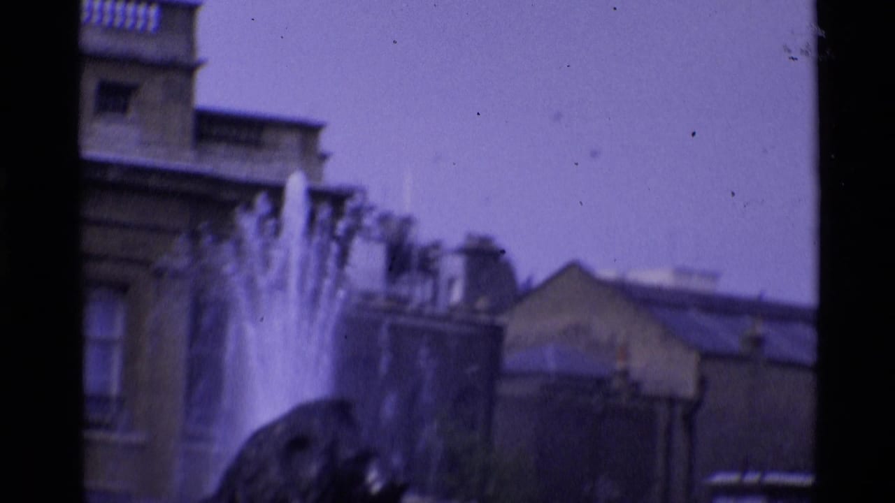 Fountain spray in front of stone buildings, seen in soft focus from a moving camera.