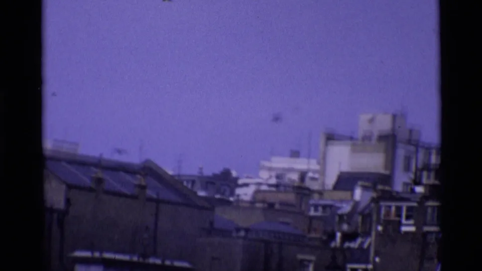1969 London rooftop view with layered building tops, chimneys, and soft purple-tinted 8mm texture.
