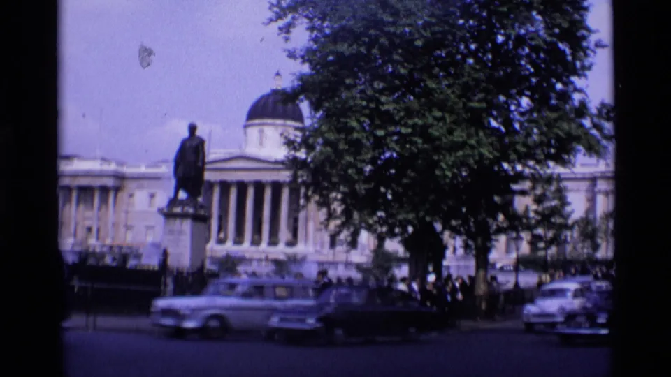 1969 London street view with a domed neoclassical building, statue, tree cover, passing cars, and pedestrians in the distance.