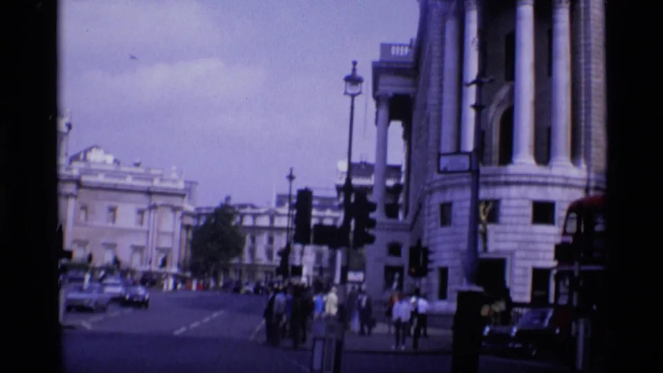 1969 London street corner seen from a handheld camera, with formal stone facades, traffic lights, cars, and pedestrians crossing or waiting.
