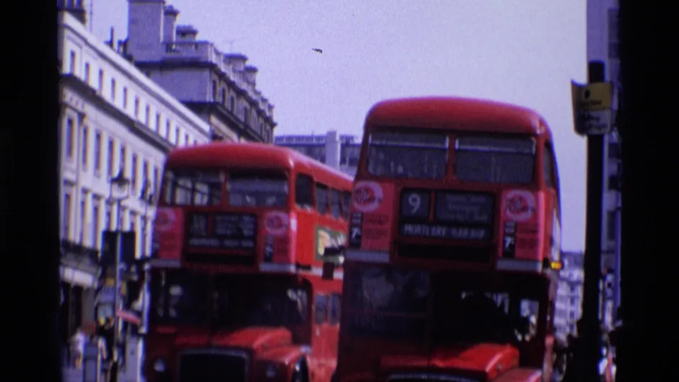 1969 London street view with two red double-decker buses, visible route signage, pedestrians, traffic signals, and multi-story building facades.
