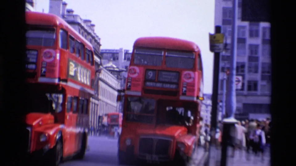 1969 London street view with red double-decker buses passing close to the camera and pedestrians near city buildings.
