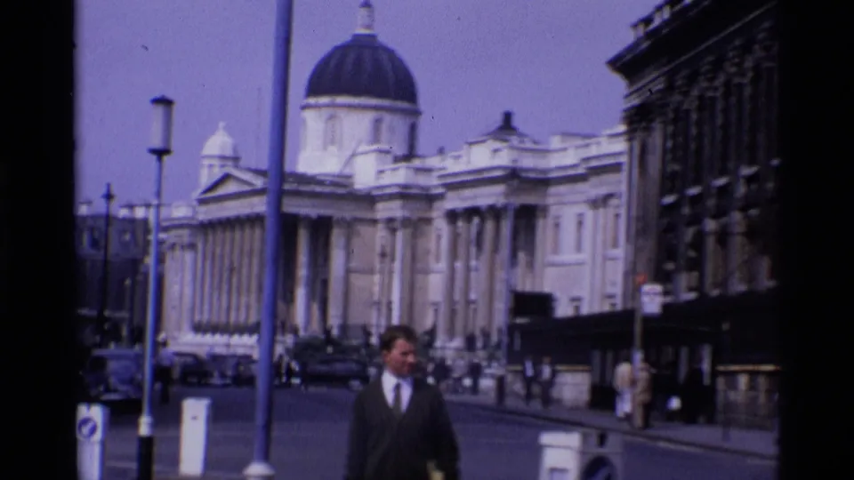 1969 London street view with a suited pedestrian in the foreground, cars, street lamps, and a domed classical building behind.