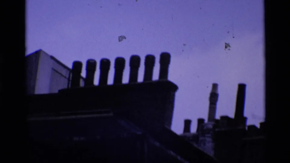 1969 London rooftop silhouette with clustered chimney pots and a purple-tinted sky in grainy 8mm footage.