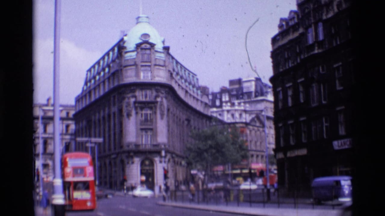 Red double-decker bus passes a corner building on a busy London street.