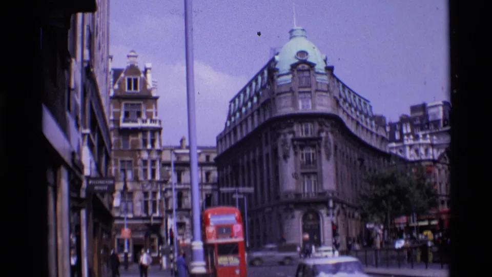 1969 London view from the pavement, showing a red bus, classic cars, pedestrians, and ornate buildings around a street corner.