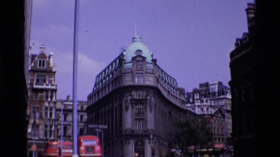 Late-1960s London street scene with a red bus, classic cars, pedestrians, and a domed stone corner building in view.