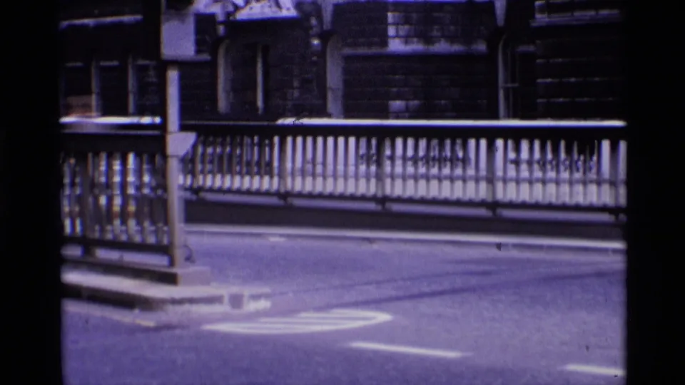 1969 London home-movie frame showing a bridge railing, pale road markings, and dark masonry building fronts beyond the street.