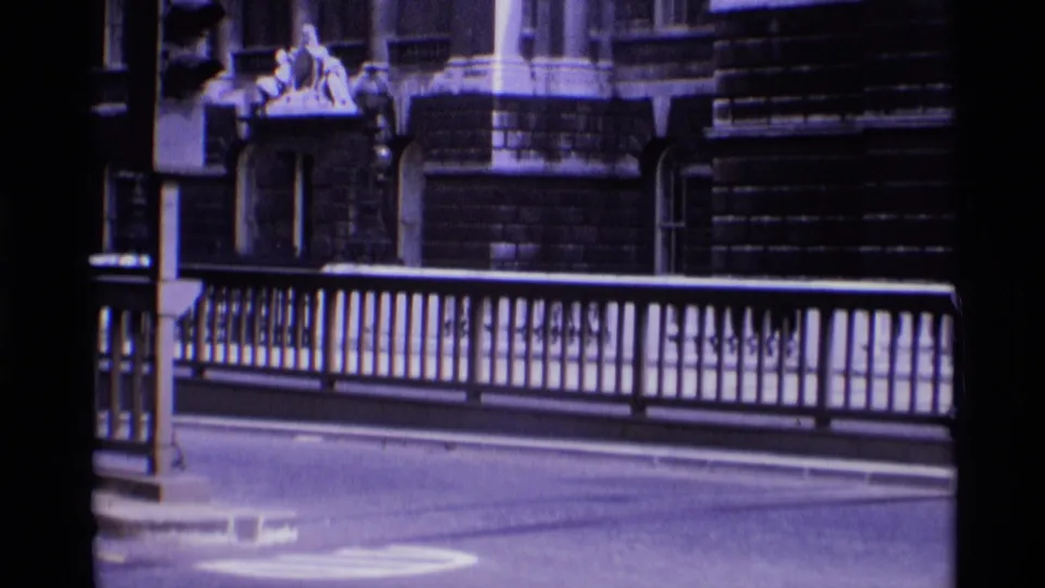 1969 London frame with metal bridge railings, quiet roadway, and an ornate building facade visible behind the street.