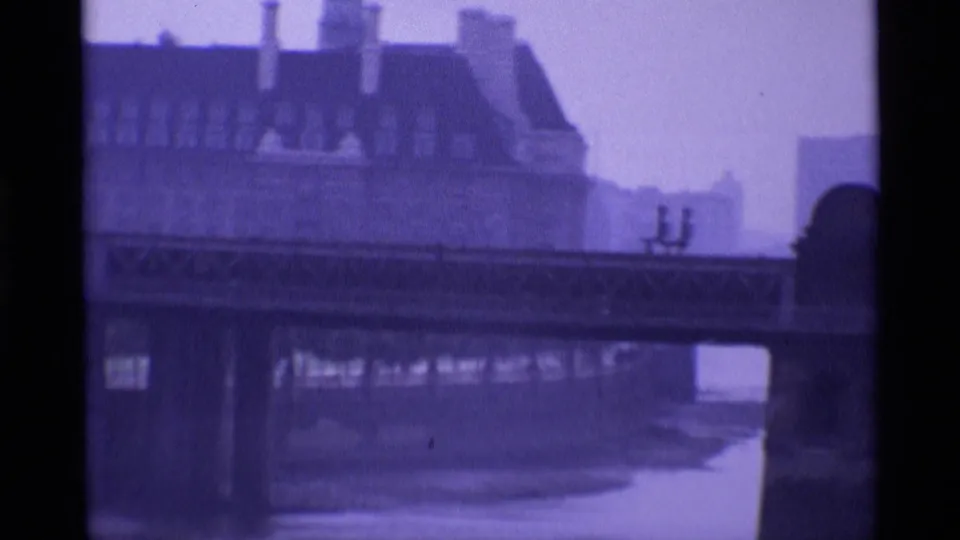 1969 London frame showing a bridge above water, a large roofline behind it, and two distant seated figures near the railing.