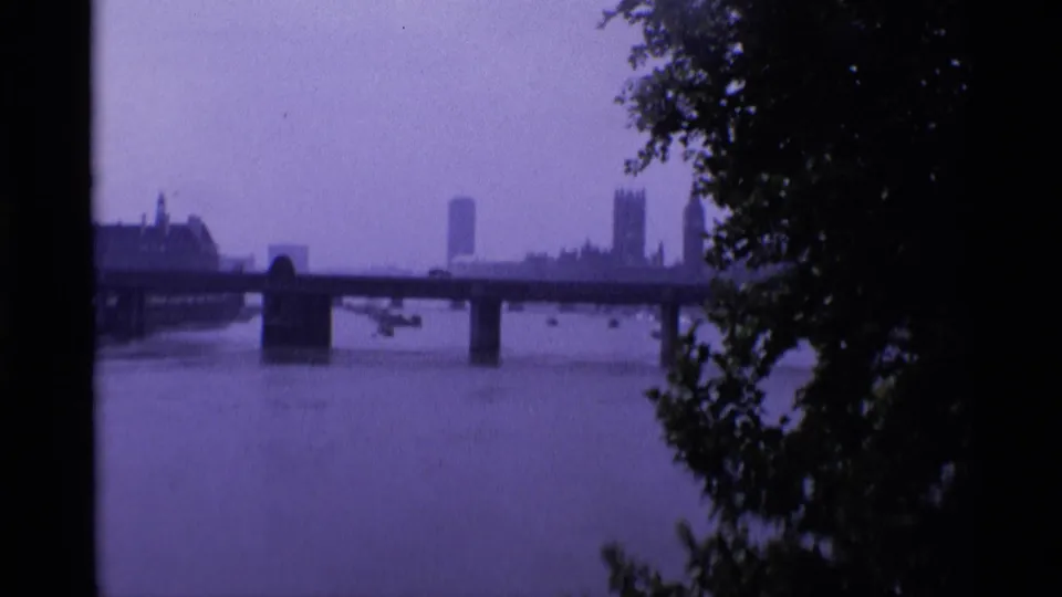 1969 London home-movie view across the River Thames toward a bridge and distant Westminster skyline, partly framed by dark trees.