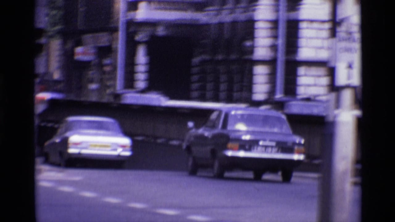 Classic cars move along a London street in 1969, with masonry buildings and soft 8mm motion blur in the frame.