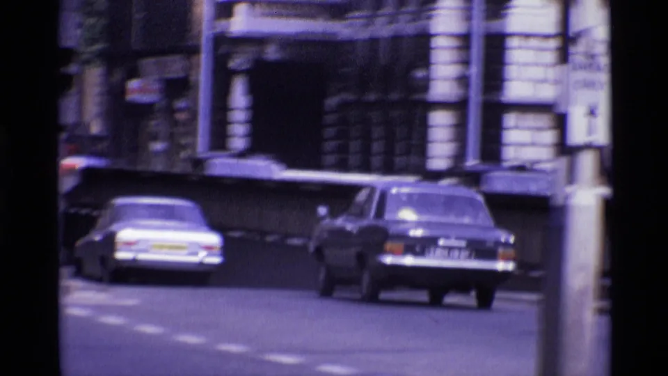 Classic cars move along a London street in 1969, with masonry buildings and soft 8mm motion blur in the frame.