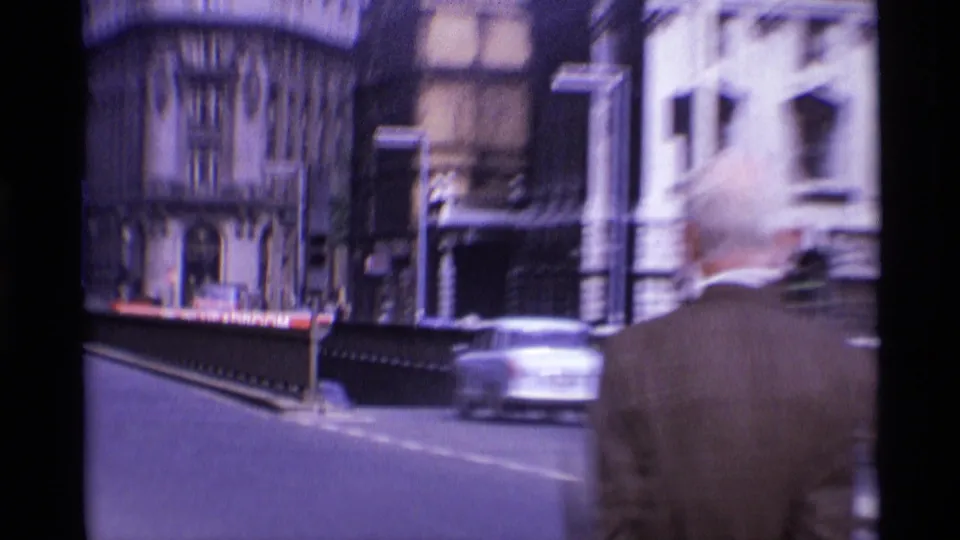 A handheld 1969 London street view shows a large stone building, passing car and partial back of an adult figure in the foreground.